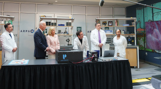 A group of people in lab coats and formal attire stands in a modern laboratory. The scientists demonstrate the MAGIC-SCAN imaging system to former President Joe Biden and Dr. Jill Biden.