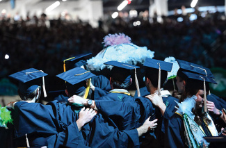 Graduates in caps and gowns stand with arms around each other, facing the audience, as one holds a decorated umbrella topped with feathers during Commencement