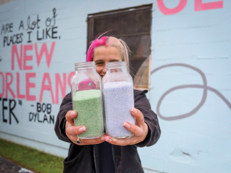 A person with pink and blonde hair holds two jars of colored sand, green and blue. They stand in front of a mural saying "New Orleans" in pink letters.