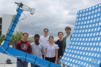 A group of six people stands beside a large, curved blue solar panel on a rooftop under a cloudy sky, conveying teamwork and innovation.