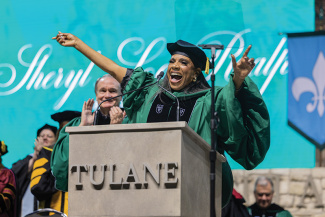 actress Sheryl Lee Ralph stands at a podium raising both arms enthusiastically while addressing the audience, with faculty members clapping in the background