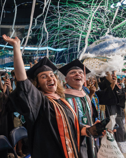 rows of graduates in cap and gown celebrate beneath blue and green streamers falling from the ceiling of the Superdome 