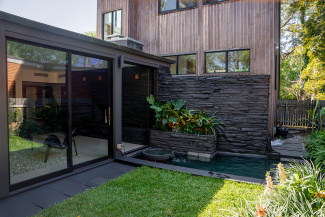 Modern backyard with a small water feature and lush greenery next to a wood-paneled house. Glass doors reflect natural light, conveying a serene atmosphere.