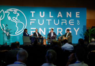 Panel discussion at "Tulane Future of Energy" event. Four people seated on stage, framed by a blue backdrop with globe graphic, addressing the audience.