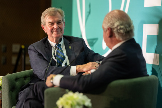 Two men in suits engage in a conversation on stage, seated in green armchairs. One smiles warmly while listening, conveying a professional yet friendly tone.