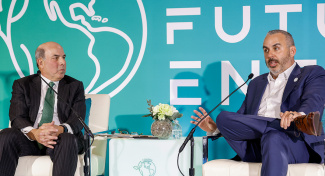 Two men in suits sit on stage in white chairs during a discussion. The atmosphere appears professional and engaged.