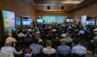 Audience seated in a conference hall at the Tulane Future Energy Forum. Large screens and banners display the event logo. Engaged attendees face a panel.