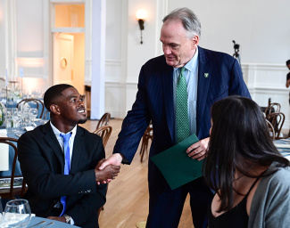 A man in a suit warmly shakes hands with another man in a formal setting, both smiling. A woman is seated nearby. The atmosphere is formal and cordial.