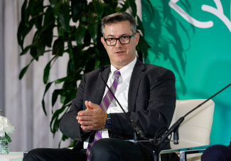 A man in a suit and purple-striped tie speaks while seated on a stage with green and white backdrop. He appears engaged and thoughtful.