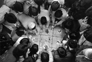 A group of people gather closely around a game board on the ground, exchanging money. The scene is busy and energetic, capturing intense focus and engagement.