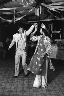 A joyful couple dances under streamers and a disco ball. The man wears a suit, while the woman wears a traditional dress with a matching headpiece. 