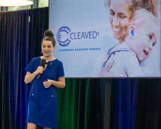 A woman in a blue dress speaks into a microphone in front of a screen displaying the Cleaved logo with an image of a smiling woman holding a child with a hearing aid.