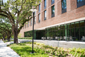 Cozy patio of a brick building with large windows, lined by tables and chairs. A path with lush greenery and tall oaks creates a serene atmosphere.