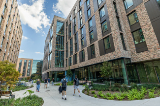 Students walk along a path beside modern brick buildings under a blue sky. The area is landscaped with greenery.