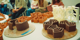 3D-printed architectural models in brown and white feature geometric designs on display tables. People in casual attire sit in the background.