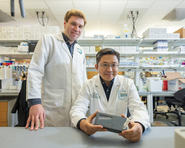 Researchers Brady Youngquist and Tony Hu in lab coats stand indoors holding a box, surrounded by laboratory equipment and medical instruments.