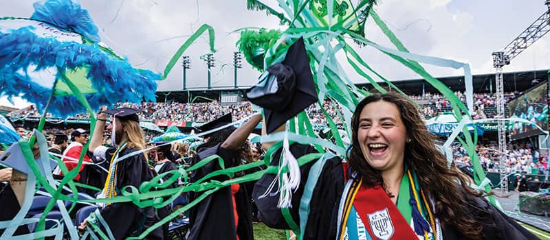 Crowd of graduates celebrate as streamers come down on a stadium field. A graduate in the foreground holds decorated umbrella.