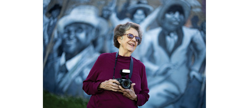 Judy Cooper holds her camera and stands in front of mural depicting second line dancers
