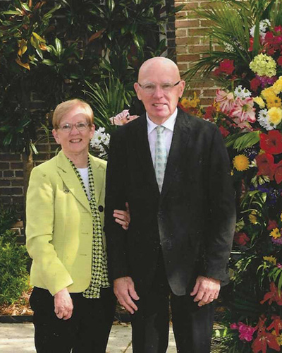 Gene and Mary Koss stand outside with flowers in the background