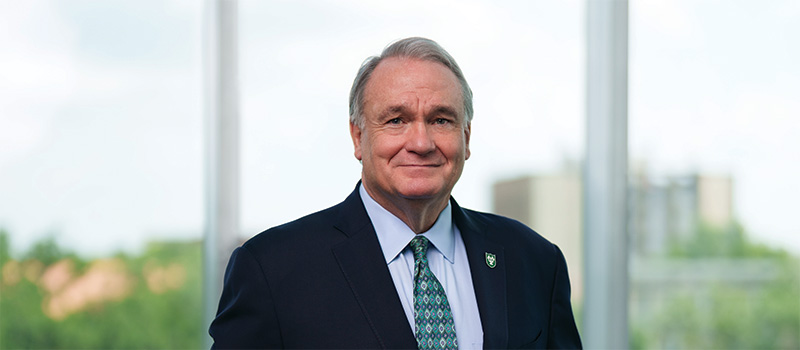 President Fitts stands in front of large windows overlooking campus buildings and treetops