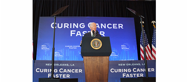 President Biden speaks from stage with signs that read Curing Cancer Faster 