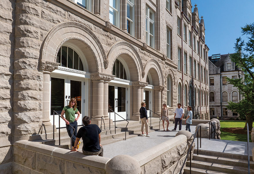 Several people gather outside a historic stone building on Tulane University's campus, chatting in small groups. The building features arched doorways, decorative stonework, and tall windows, with a bright blue sky and green lawn in the background.