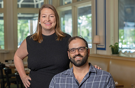Restaurant owners Alon and Emily Shaya pose together inside a bright, stylish dining space, Saba. Emily, wearing a sleeveless black dress, stands with one hand on her hip and the other resting on Alon’s shoulder. Alon, seated and wearing glasses and a blue plaid shirt, smiles toward the camera. Large windows and tables are visible in the background.