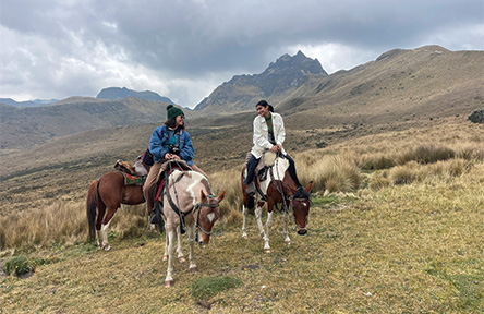 Two people ride horses on a grassy mountain landscape under a cloudy sky. They seem relaxed, engaged in conversation, with rugged peaks in the background.