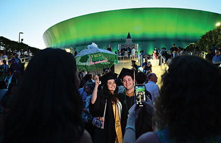 graduates in cap and gown pose for a photo outside a green-lit Superdome at dusk, surrounded by a crowd of people