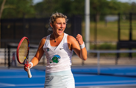 A female tennis player wearing a white outfit passionately celebrates with a fist pump on an outdoor court, holding a red racket.