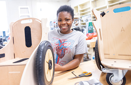 A student smiles while assembling a wooden mobility device at a workshop. The space is bright with tools and materials scattered around, conveying creativity.