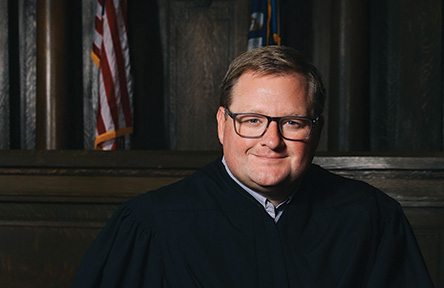 Justice Cade Cole in black judicial robes sits in a formal courtroom setting, wearing glasses and holding a book.