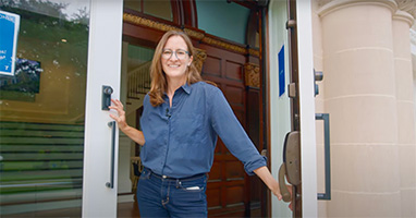 A smiling person with long hair and glasses holds open a glass door, wearing a blue shirt and jeans. The background features wooden paneling and a stone column, suggesting a welcoming entrance.