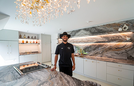 A smiling man in a cowboy hat stands in a modern kitchen with marble countertops and backsplash. Soft lighting creates a warm, inviting atmosphere.