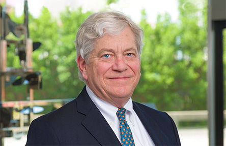 A man with white hair smiles warmly, wearing a dark suit with a blue polka dot tie in front of a window showing lush greenery outside.