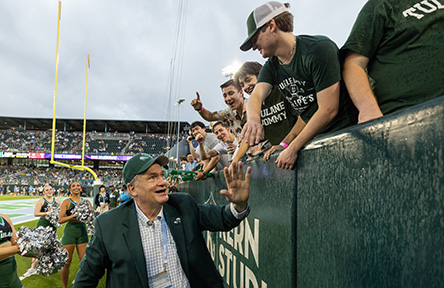 A man in a suit and cap high-fives enthusiastic fans at a football game. Cheerleaders stand nearby. The atmosphere is lively and celebratory.