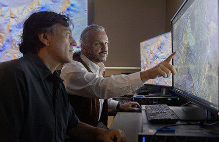 Two men analyze a detailed map on a large monitor. One points at the screen, focusing intently. The room is dimly lit, suggesting concentration.