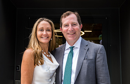 A smiling woman in a white dress and a man in a gray suit with a teal tie stand closely together, posing happily in front of a doorway.