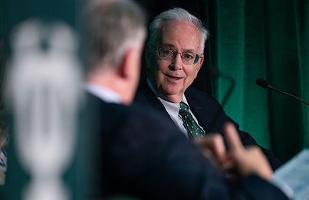A man in glasses and a suit speaks at a panel, looking engaged, with a microphone nearby. A blurred person and green curtain are in the foreground.