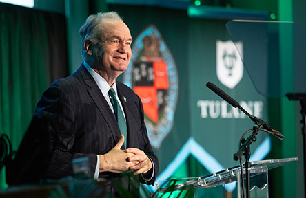 A man in a suit and tie speaks at a podium with a microphone. Behind him is a Tulane University banner. The atmosphere is professional and formal.