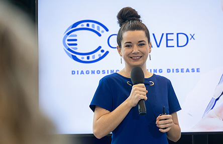 Smiling woman in blue dress holding a microphone, presenting in front of a screen with logo. 