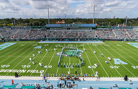 Aerial view of a football stadium with a marching band forming a large letter "B" shape on the field. Spectators fill the stands, and the atmosphere is lively.