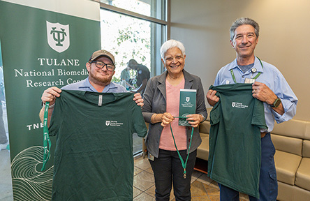 Three people stand smiling, holding green Tulane T-shirts. A Tulane National Biomedical Research Center banner is behind them, conveying a celebratory mood.