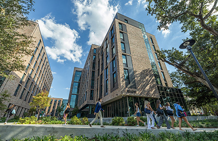 A group of students walks past modern, multi-story brick buildings under a blue sky with scattered clouds. The scene is lively and academic.