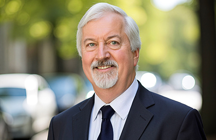 Bill Templeton in a suit smiling outdoors, with blurred trees and cars in the background. The scene conveys a professional and approachable tone.