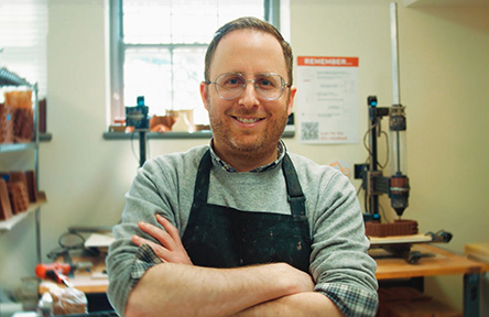 Smiling person with glasses and beard, arms crossed, wearing a black apron in a workshop. Shelves and tools create a cozy, industrious atmosphere.