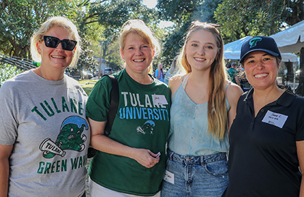 Four people smiling outdoors, wearing casual Tulane University apparel. Sunlight filters through trees, creating a warm, cheerful atmosphere at an event.