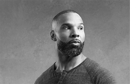 A black and white photo shows a man with a beard gazing to the side. He wears a dark, textured shirt, set against a plain background, conveying thoughtfulness.
