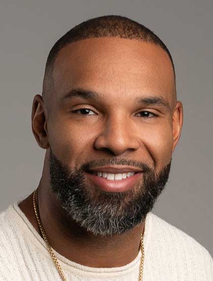 A man with a short beard and a warm smile is facing the camera. He wears a light-colored shirt and gold chain, set against a neutral gray background.