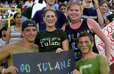 Fans in a stadium cheer energetically, holding a sign that reads "Go Tulane Green Wave." Faces are painted green, expressing school spirit and excitement.